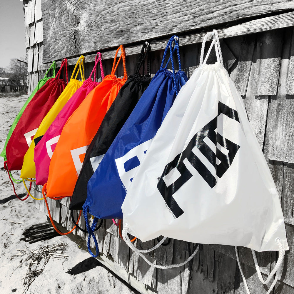 A collection of colorful cinch sacks with drawstrings, hanging on a wooden fence. They come in various colors including pink, yellow, orange, blue, and white, with PTOWN logo printed on the front in black.